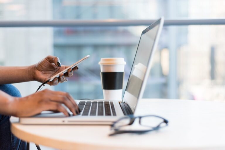 Person working on laptop, holding phone, coffee nearby. Remote work, digital nomad, productivity.