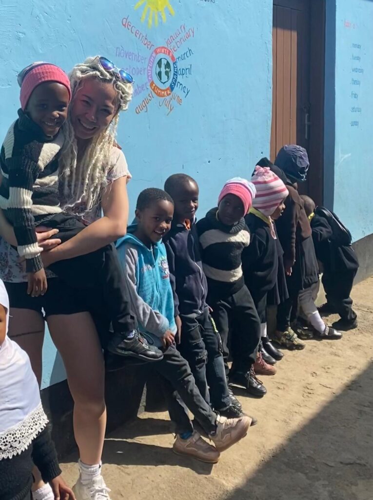 Group of children with woman, smiling outside blue wall.